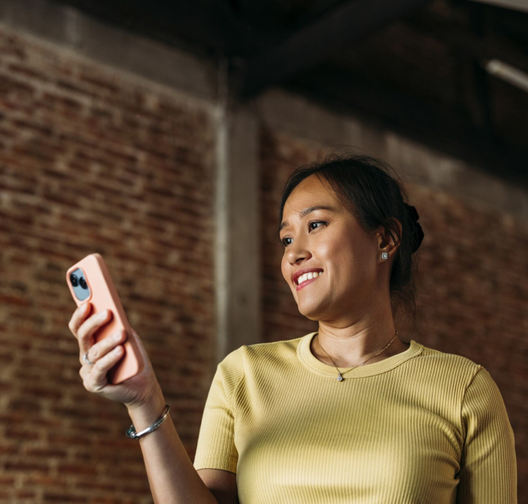Woman in yellow t-shirt standing by brick wall and smiling at phone in hand