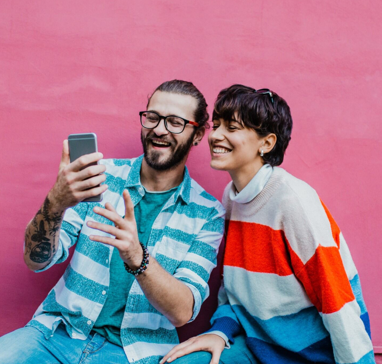 Girl with short hair sitting on ground next to man with glasses showing her something on phone, both sitting in front of a pink wall