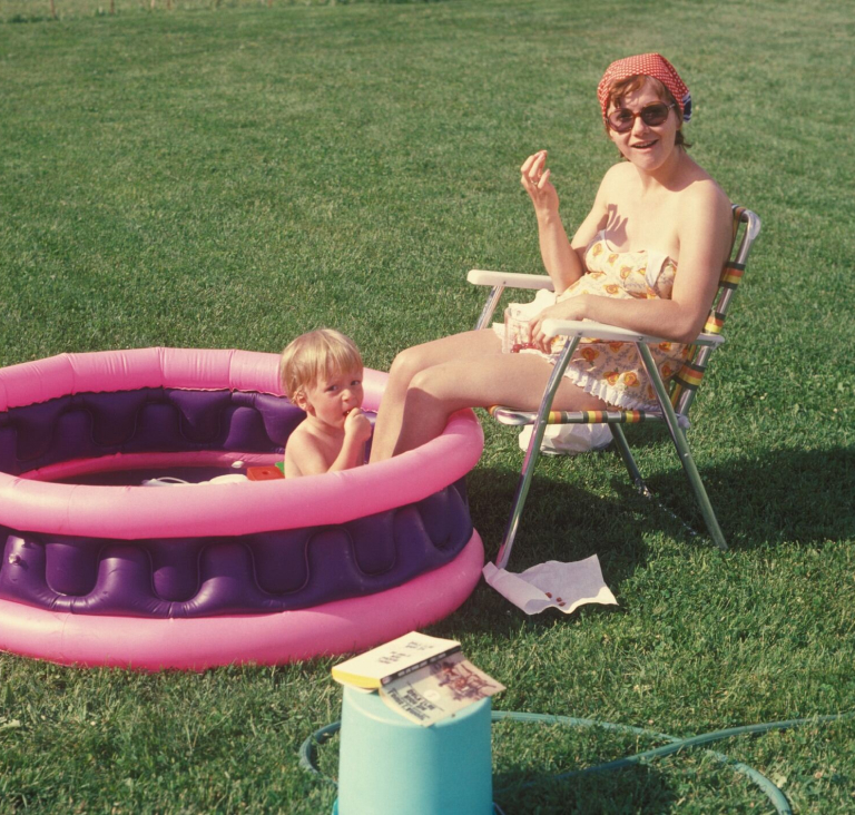 Woman sitting in lawn chair in garden, while child places in pink and purple inflatable pool