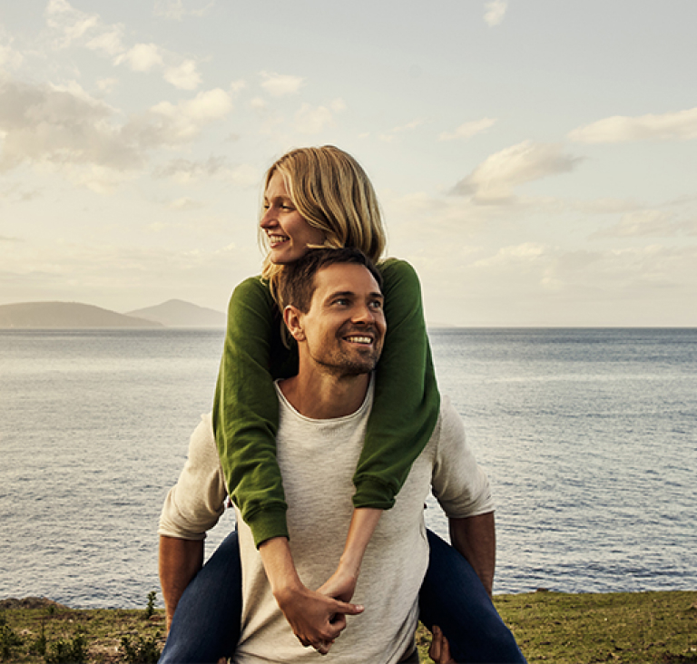 Woman piggy backing man, standing on grassy land in front of ocean