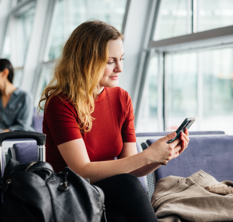 Woman in red top holding phone and sitting in airport with suitcase and bags