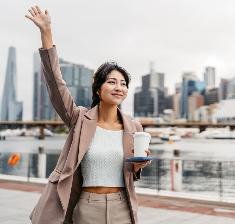 Woman walking alongside river holding coffee cup in one hand and raising her other arm in the air