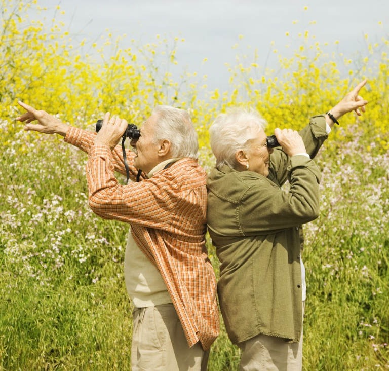 Couple standing back-to-back in a field, holding binoculars and pointing in different directions