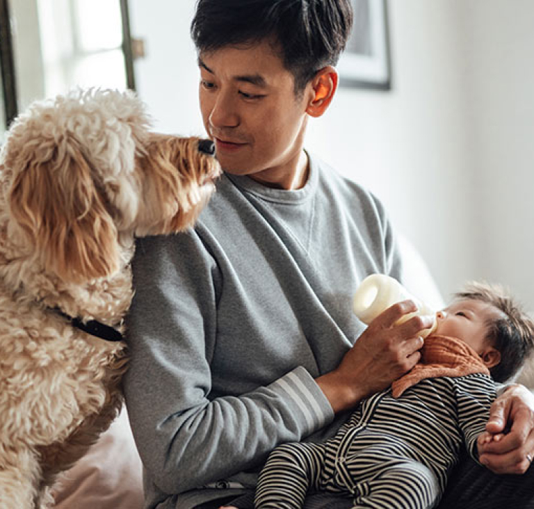 Man sitting on sofa feeding baby from bottle, with dog sitting by his side on the couch