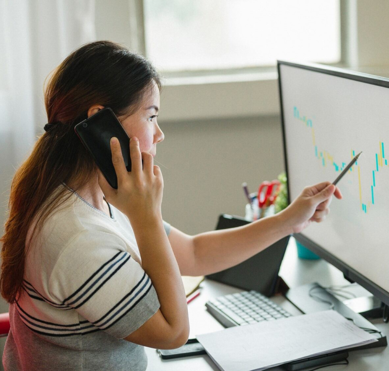 Girl sitting at desk, holding phone up to ear while pointing pen at screen with graph on it