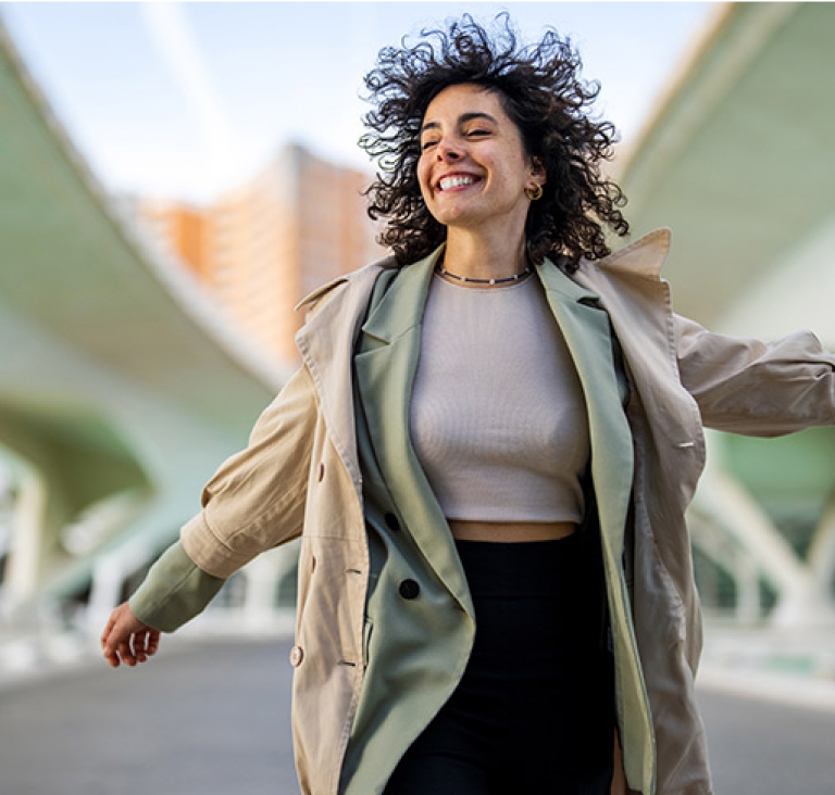 Woman wearing coat and walking under two bridges, smiling with arms slightly out