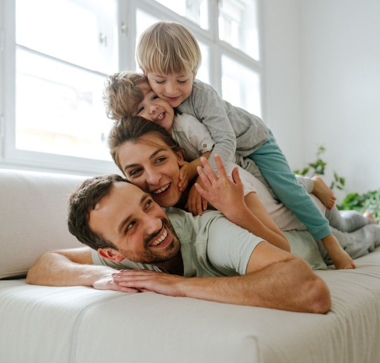 Family of two parents and two young children lying on top of each other on a sofa in their living room