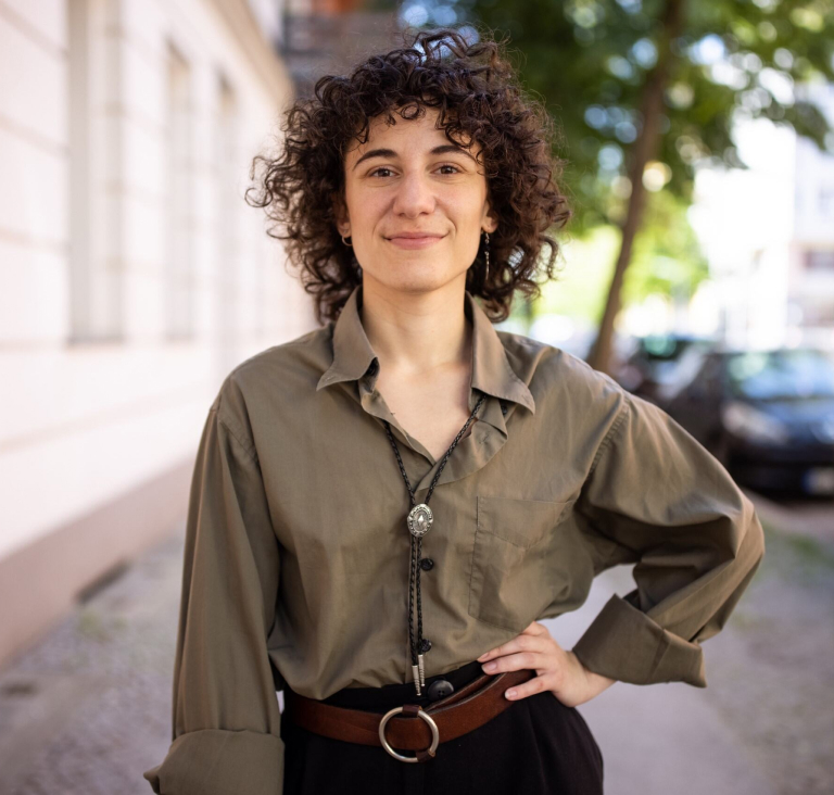 Woman in khaki shirt standing on street pavement with hand on hip