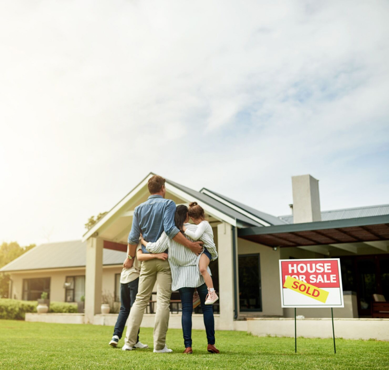Couple with two kids standing on front lawn facing house with sold sign next to them