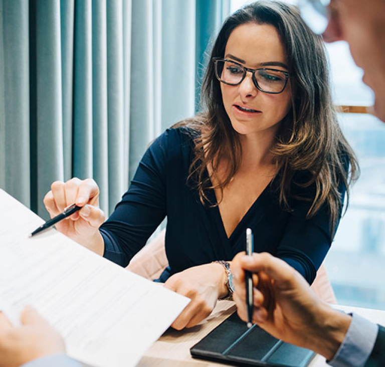 Woman seated at table wearing glasses and pointing with pen at document that man is holding
