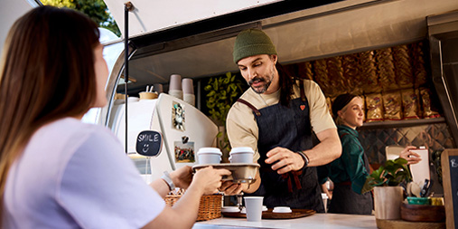 A barista working in a coffee truck hands two takeaway coffees to a customer