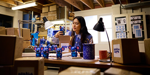 A woman packaging handmade products at a workshop table surrounded by boxes