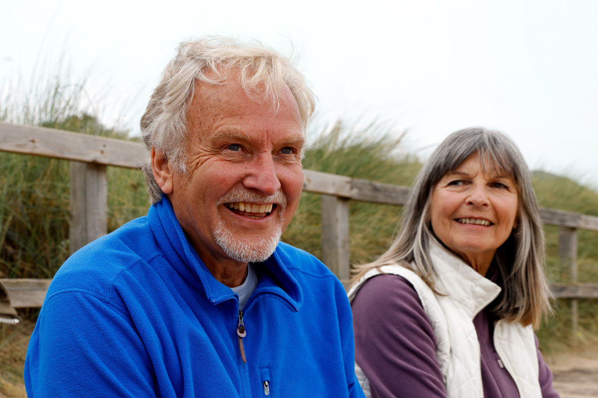 Elderly man, smiling and wearing a blue jumper, sitting with an elderly woman.