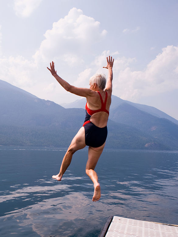 Person wearing a red and black swimsuit leaping into a lake, arms raised, with mountain ranges and cloudy skies in the background.