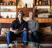 Two people sitting together on a couch with shelves and framed photos behind them.