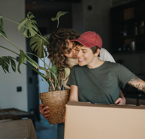 Two people are inside a home, one person holds a large potted green plant and the other is standing close, with their arm around the first person. They are surrounded by moving boxes, suggesting a moving or unpacking scene. 