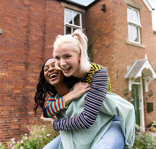 A woman gives her friend a piggyback in front of a red brick house