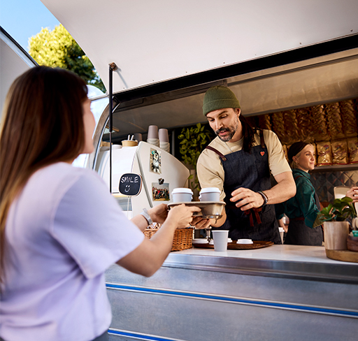 A barista wearing an apron and a beanie in a coffe truck hands two takeaway coffees to a customer