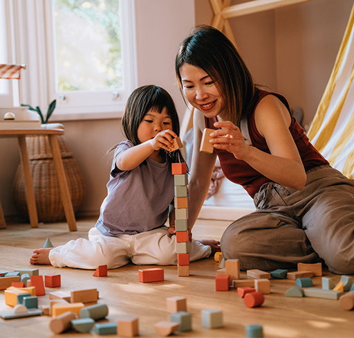 A woman and a young child sit on the floor of a cozy room, building a tall tower together with colourful wooden blocks.