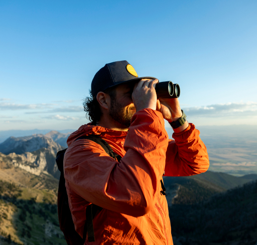 Person in an orange jacket uses binoculars on a mountain ridge overlooking a vast landscape.