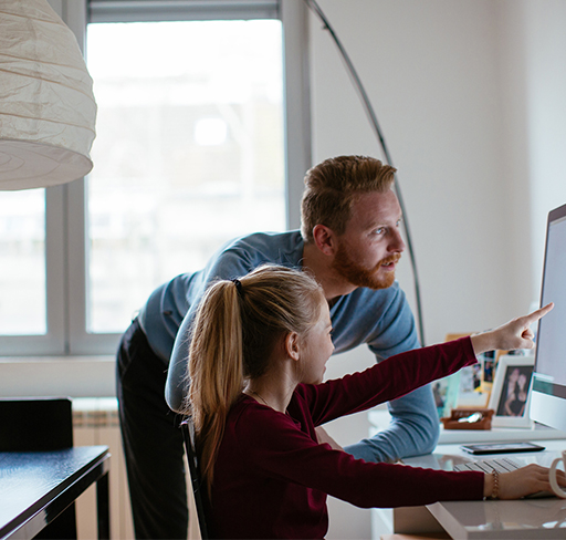 A young girl sits at a desk in a modern home office setting, pointing at a computer screen. Beside her, a man leans over the desk, observing the screen.