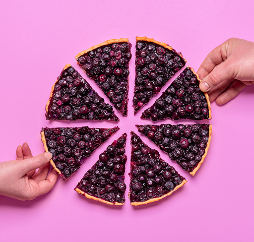 Two hands reach for slices of a blueberry pie that has been cut into eight pieces and arranged in a circle on a pink background.