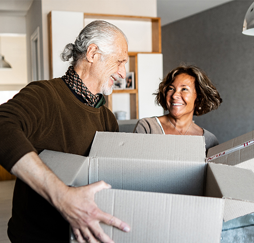 An older man with gray hair in a ponytail, wearing a brown sweater and scarf, handles a large cardboard box. Beside him is a woman with short, wavy hair in a casual top. They are in a modern, well-lit living space with contemporary decor.