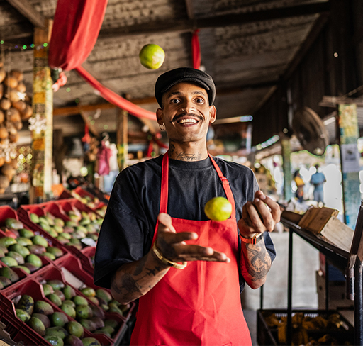 A person wearing a red apron is juggling limes in a rustic fruit market, surrounded by stacks of fresh produce and hanging decorations.