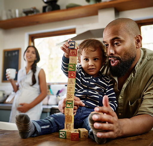 A child builds a tall tower with alphabet blocks on a kitchen counter, assisted by their father. In the background, a woman holds a cup near the sink with sunlight streaming through the windows.