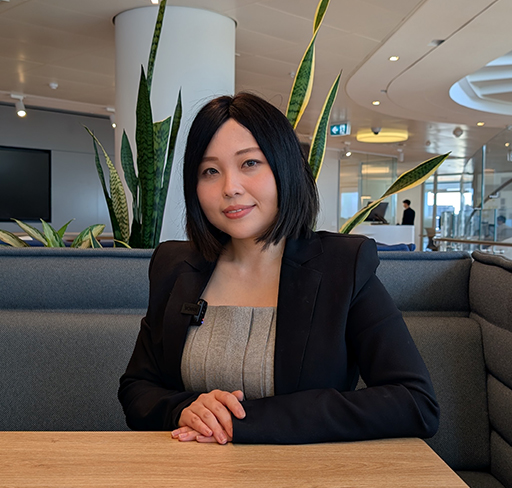 A person in business attire sits at a table in a modern office lounge with plants and glass architecture in the background.