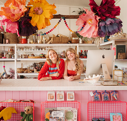 Two women stand behind a counter in a vibrant and colourful shop decorated with large paper flowers and garlands, surrounded by shelves filled with various art and craft products.