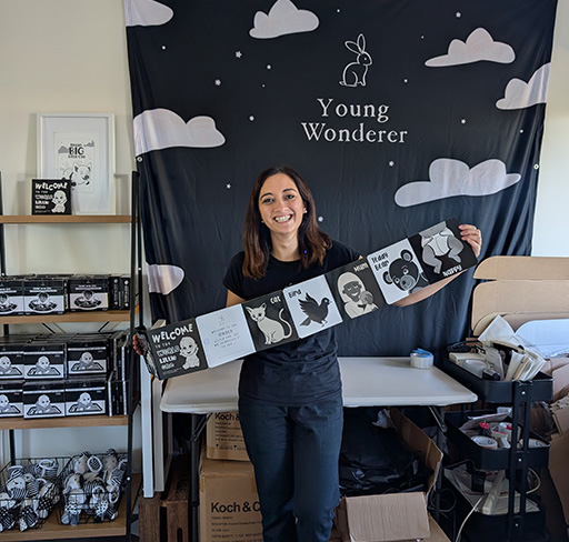 A baby product business owner holds a black and white illustrated baby book in front of a backdrop with the words "Young Wonderer," surrounded by shelves filled with baby books and boxes.