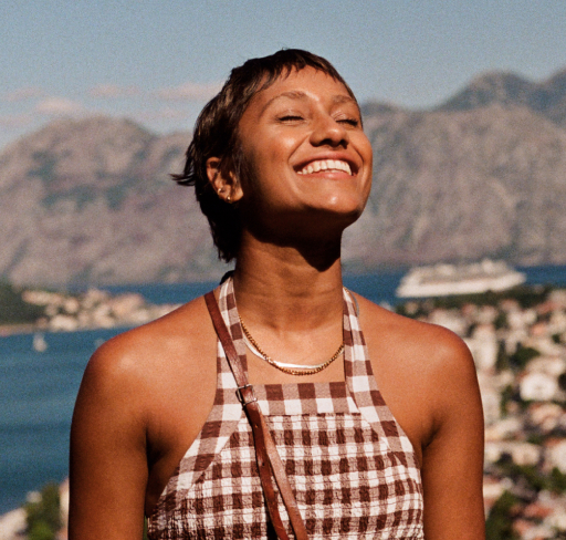 A woman with cropped hair enjoys the sunshine on holiday wearing a sleeveless checkered top standing outdoors with a coastal town, blue water and mountain landscape in the background.