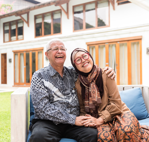 An elderly couple dressed in batik patterns sit closely on an outdoor bench in front of a house, smiling at the camera with arms around each other and hands clasped.