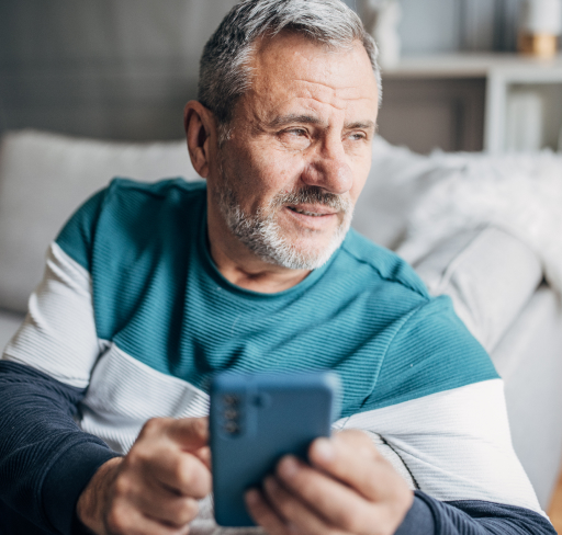 A middle‑aged man sits on a sofa at home, holding and looking down at a smartphone while wearing a casual long‑sleeve top.