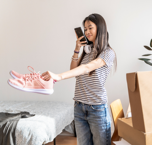 A woman photographs a pair of pink running shoes for an online listing while holding them out beside a bed and shopping bags in a home setting.