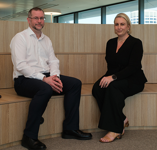 A man and a woman in business attire sit on tiered wooden seating in a modern office space with large windows.