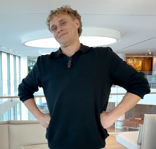 A person with long brown hair, wearing a light blue striped shirt and dark trousers, sits at a desk in a modern office space and types on a laptop, with a large monitor and glass walls in the background.