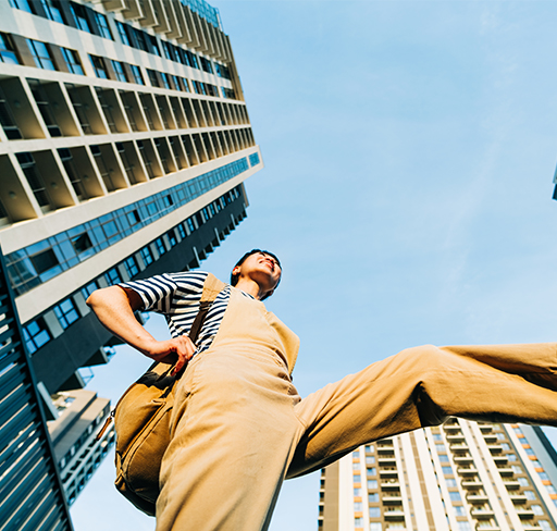 A person wearing tan overalls, a striped shirt, and white sneakers is captured from a low angle, stepping forward confidently among tall, modern apartment buildings under a blue sky.