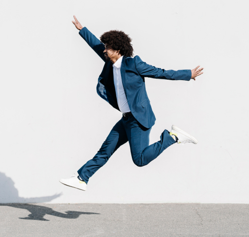 A person in a blue suit and white sneakers is captured mid-air in a side leap against a plain white wall, with arms outstretched and a shadow cast on the ground below.