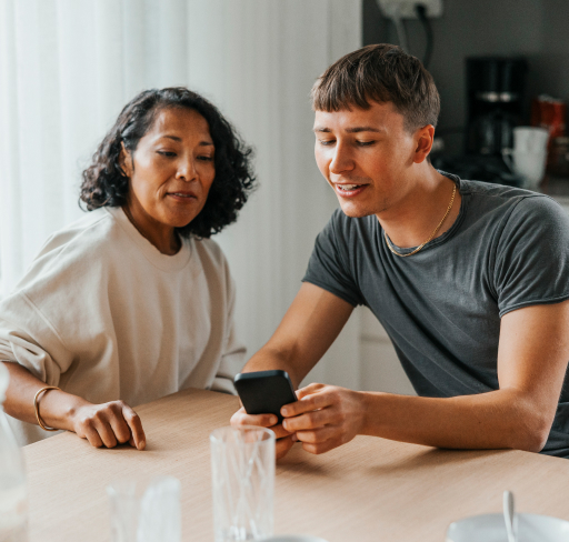 A woman and a younger man sit at a kitchen table and look at the man's phone.