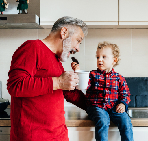 An adult man wearing a red top stands in a modern kitchen holding a mug while facing a small child in a red plaid shirt sitting on a countertop, with a kettle and cabinets visible in the background.