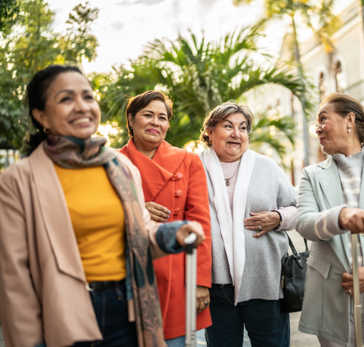 Four women walk together through a leafy outdoor garden, dressed in smart-casual clothing and enjoying a social outing in a natural setting.
