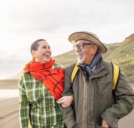 Copilot said:  Two adults wearing outdoor clothing and scarves walk arm in arm along a beachside path, with coastal hills and the ocean visible in the background.