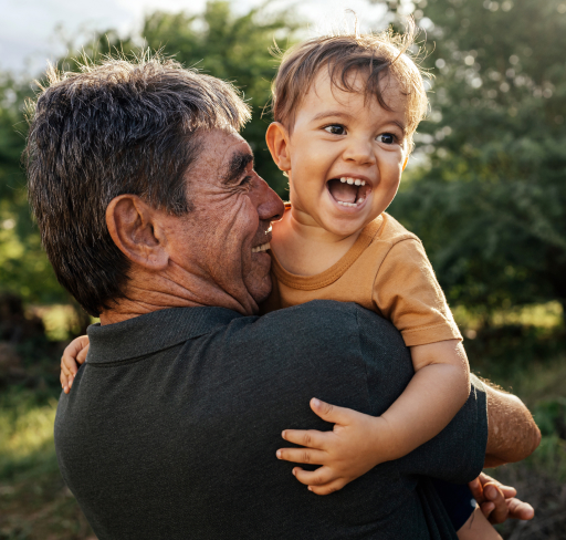 An adult man carrying a young child in an outdoor setting, with trees and soft natural light behind them.