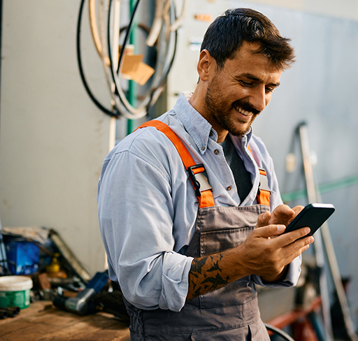 A tradesperson wearing grey overalls with orange straps and a light blue shirt stands in a workshop, holding and using a smartphone. Tools and cables are visible in the background, and the person has a tattoo on their forearm.
