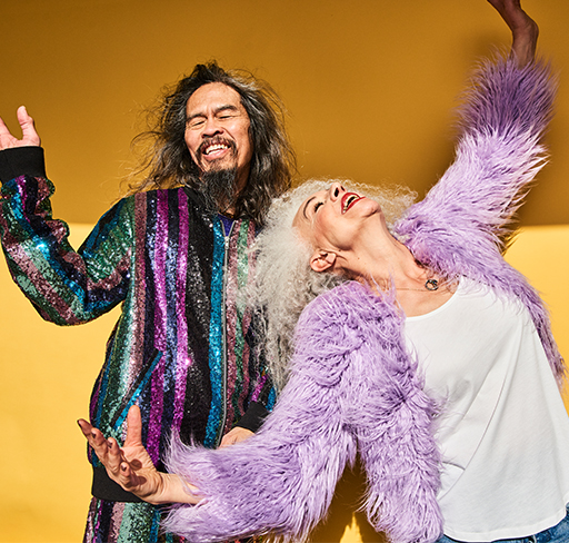 Two people pose playfully against a mustard-yellow backdrop, one wearing a multicoloured sequined jacket and one wearing a fluffy purple coat over a white top, with their arms raised in a carefree gesture.