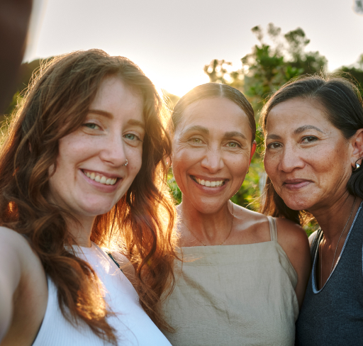 Three women of different ages gathered together outdoors, with soft evening light and greenery in the background
