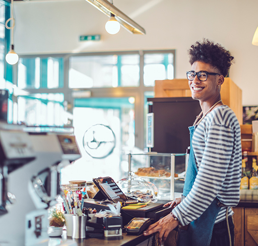 A person wearing a striped long-sleeve shirt and a blue apron stands behind the counter at a modern café. 