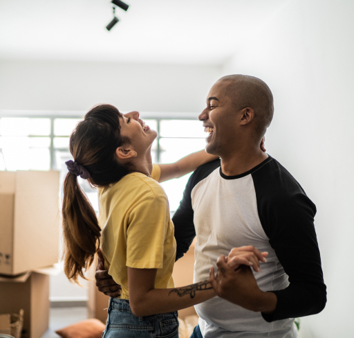 Happy young couple dancing while moving house, with moving boxes in the background.